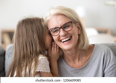 Cute small granddaughter whisper something in granny ear, grandmother and grandchild share secrets having sincere talk at home, grandma and little girl have fun at home together laughing - Powered by Shutterstock