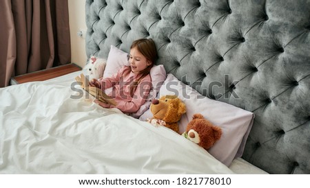 A cute small happy girl lying with teddybears in a huge bed covered with blanket smiling and reading book aloud. relaxing in a big bright bedroom at home