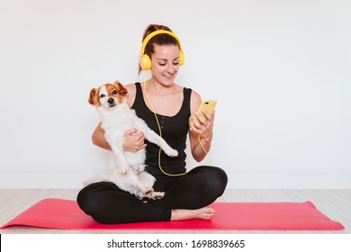 Cute Small Jack Russell Dog Doing Yoga On A Mat At Home With Her Owner. Young Woman Listening To Music On Yellow Mobile Phone And Headset.Healthy Lifestyle Indoors