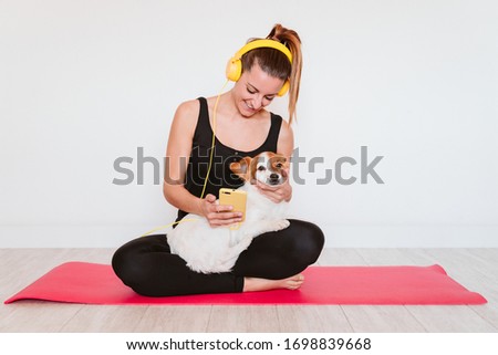 cute small jack russell dog doing yoga on a mat at home with her owner. Young woman listening to music on yellow mobile phone and headset.Healthy lifestyle indoors