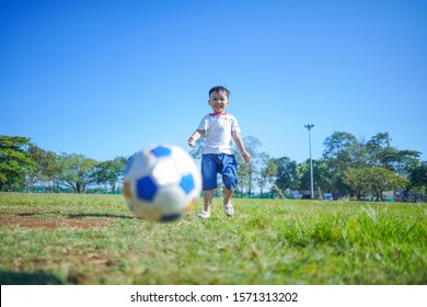 Cute Small Kid Playing Soccer On Soccer Ground During Clear Blue Sky, Little Child Smiling And Kicking Soccer Ball On Green Grass Field Alone, Boy Kick The Ball Soaring Above The Football Ground