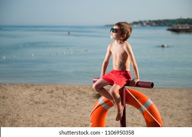 Cute Small Kid In Sunglasses Sitting On Beach With Lifebuoy Outdoor Summer Like Baywatch Movie Heroes