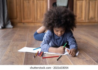 Cute Small Teen African American Girl Child Lying On Warm Wooden Kitchen Floor Drawing Painting In Album Alone. Little Teenage Ethnic Biracial Kid Have Fun Rest At Home Engaged In Art Hobby Activity.