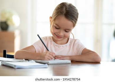 Cute smart little 7s girl child sit at table at home write in exercise book prepare homework alone. Happy small kid do task assignment, handwrite in notebook, study for school. Education concept.