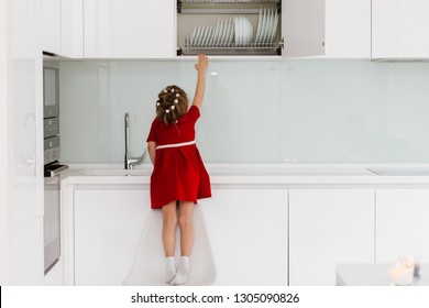 Cute Smart Little Caucasian Girl In Casual Red Dress Climbed Onto A Chair And Trying To Reach Dishes In A Closet In A Modern Minimalist Kitchen