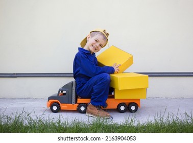 cute smiling 6 year old boy in uniform sits on a big toy car - a truck with many cardboard boxes. Parcel delivery, little postman, truck driver. Positive, joy, games for boys. work like a dad