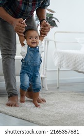 Cute Smiling African Baby Girl Learning To Walk With Father Support And Holding Her Hands. Happy Little Toddler Child Taking First Steps With Parents Help At Bedroom At Home With Love And Care