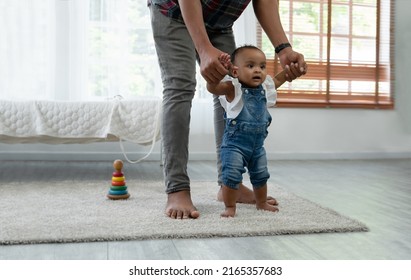 Cute Smiling African Baby Girl Learning To Walk With Father Support And Holding Her Hands. Little Toddler Child Taking First Steps With Parents Help At Bedroom At Home With Love And Care