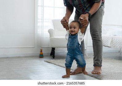 Cute Smiling African Baby Girl Learning To Walk With Father Support And Holding Her Hands. Happy Little Toddler Child Taking First Steps With Parents Help At Bedroom At Home With Love And Care