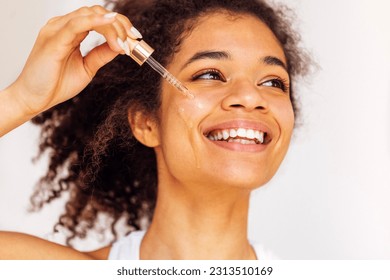 Cute smiling african young woman applies moisturizing serum from pipette to her cheek. Lovely charming mixed race girl taking care of her facial skin. Cosmetics and beauty concept.
