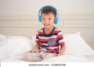 Cute Smiling Asian 3 - 4 Year Old Toddler Boy Child Wearing Striped T Shirt Listening To Music In Headphones & Singing In Bed. Little Kid Listening Loud Music In Wireless Earphones And Dancing At Home