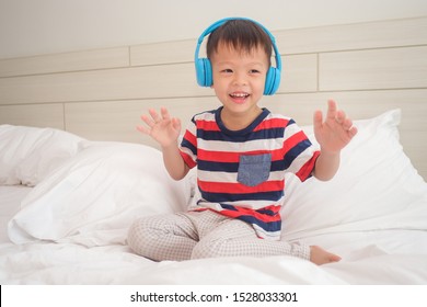 Cute Smiling Asian 3 - 4 Year Old Toddler Boy Child Wearing Striped T Shirt Listening To Music In Headphones & Singing In Bed. Little Kid Listening Loud Music In Wireless Earphones And Dancing At Home