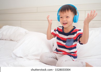 Cute Smiling Asian 3 - 4 Year Old Toddler Boy Child Wearing Striped T Shirt Listening To Music In Headphones & Singing In Bed. Little Kid Listening Loud Music In Wireless Earphones And Dancing At Home