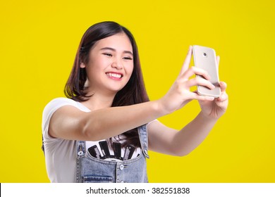 Cute smiling Asian teenage girl taking self shot photo with her phone camera, on bright yellow background