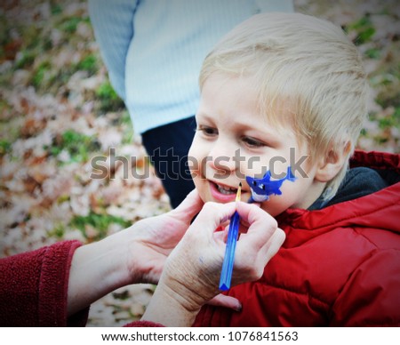 Cute smiling blond little boy in red coat getting his face painted a blue shark by face painting artist. 