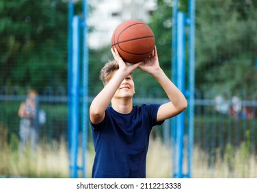 Cute Smiling Boy In Blue T Shirt Plays Basketball On City Playground. Active Teen Enjoying Outdoor Game With Orange Ball. Hobby, Active Lifestyle, Sport For Kids, Teenagers