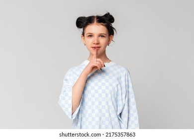 Cute Smiling Brunette Teen Girl Showing Shhh Secret Hush Gesture, Shushing, Stands In Trendy T Shirt Over Light Grey Background