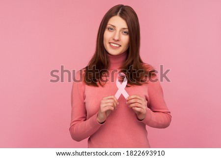 Cute smiling brunette woman in pink sweater holding pink ribbon near her chest supporting another women, international symbol of breast cancer awareness. Indoor studio shot isolated on pink background