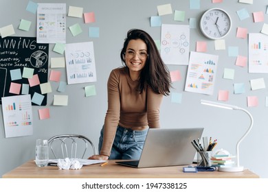 Cute, smiling business girl with glasses in the office leaning on the desk. On a gray background pasted stickers, graphics, work plans.