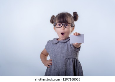 Cute Smiling Child With Glasses Holding Discount White Card In Her Hands. Kid With Credit Card. Little Girl Showing Empty Blank Paper Note, Copy Space. Isolated On Blue Background 