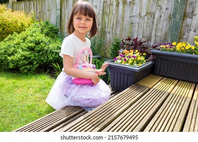 A Cute Smiling Child Is Playing In The Backyard With Small Pink Bag. Sunny Day. Good Spring Weather. Typical English Backyard