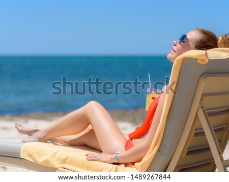 Cute, smiling European girl in sunglasses is laying on sunbed near sea. She is holding cocktail and enjoying her summer vacations.