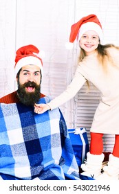 Cute Smiling Funny Blonde Girl In Beige Dress Pulls Long Beard Of Handsome Bearded Man In Blue Checkered Plaid And Christmas Red Hat On White Studio Background