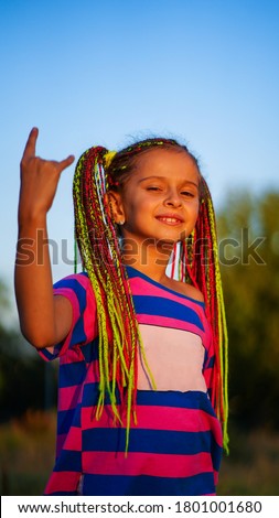 Cute smiling girl with bunched colored dreadlocks raising hand with two fingers on sunset