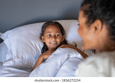Cute smiling girl lying under blanket in bed wishing good night to mother. Happy lovely daughter talking with mom at bedtime. African mother putting indian child to sleep in comfortable bed.