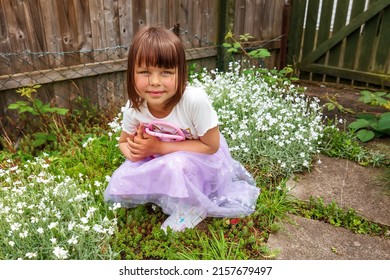 A Cute Smiling Girl Is Playing In The Backyard With Small Pink Bag. Sunny Day. Good Spring Weather. Typical English Backyard