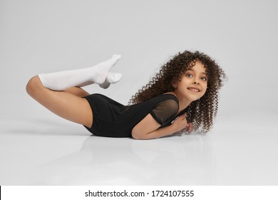Cute smiling girl in sportswear and knee socks demonstraiting boat exercise, isolated on gray background. Little female professional gymnast with curly hair showing flexibility, looking at camera.