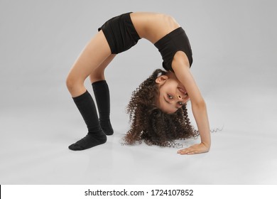Cute smiling girl in sportswear and knee socks demonstraiting bridge exercise, isolated on gray studio background. Little female professional gymnast with curly hair showing flexibility, training.