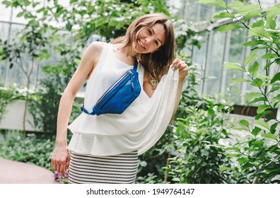 Cute Smiling Girl Walking In The Botanical Garden. The Girl Is Wearing A White Blouse And A Blue Fanny Pack.