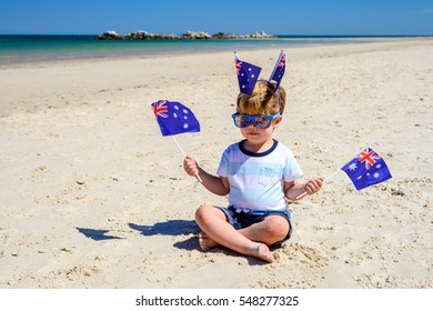 Cute Smiling Kid With Australian Flags Sitting On The Sand At The Beach On Australia Day