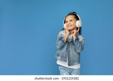 Cute Smiling Little Child Girl In White Headphones And Denim Listening To Music And Dancing On Blue Background. Copy Space