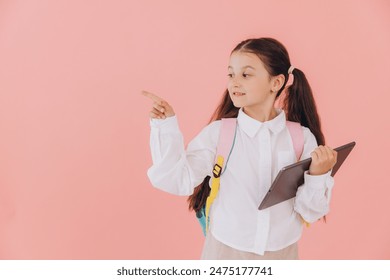 Cute smiling little girl in school uniform with backpack holding tablet and pointing something with finger on pink background - Powered by Shutterstock