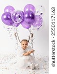 Cute smiling little girl in a white princess dress and air balloons tied to pigtails has fun with confetti on white background. Birthday party celebration. Copy space. High quality photo