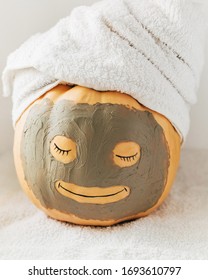 A Cute Smiling Pumpkin With Facial Clay Mask And White Towel On Light Background. Spa, Relax, Health And Beauty, Skin Care Concept. Halloween