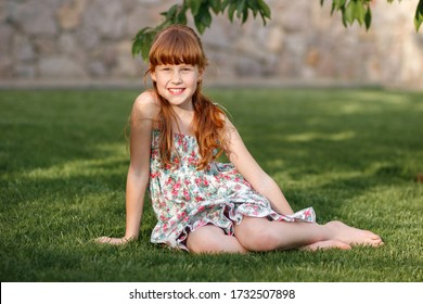 Cute smiling red-haired girl with freckles 10 years old sits carefree on the green grass in the sun in a summer dress in the open air.