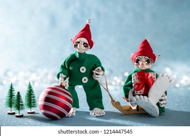 Cute Smiling Santas Helper Elves Holding Christmas Bauble And A Christmas Gift. North Pole Christmas Scene. Santas Workshop. Elves At Work. 