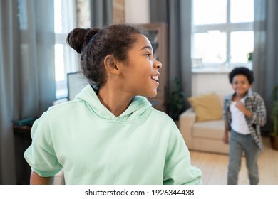 Cute Smiling Schoolgirl In Hoodie Turning Back To Take A Look At Her Brother Running After Her During Play In Living-room On Weekend