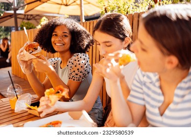 Cute smiling teenage girls are sitting in open air cafe and eating fast food. Cheerful happy female friends of different nationalities snacking on burgers and pizza outdoors and having fun together. - Powered by Shutterstock