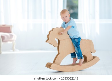 Cute, Smiling, White, Two Years Old Boy In Blue T-shirt And Jeans Rocking On Wooden Handmade Horse. Little Child Having Fun With Pony Toy. Concept Of Early Childhood Education, Happy Family, Parenting