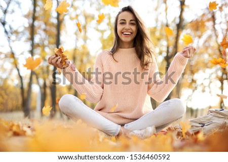 Cute smiling woman woman in autumn park with fall colorful background, sitting on blanket, throwing autumn fall leaves, enjoying warm sunny weather. Fall season concept. Outdoor lifestyle portrait.