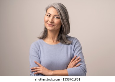 Cute Smiling Woman With Grey Hair Bends Her Head, Looks At Camera While Having Her Arms Crossed. Beautiful Middle-Aged Asian Woman With Crossed Arms. Portrait. Studio Photoshoot