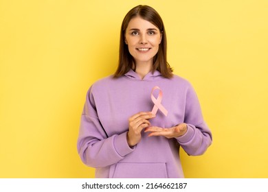 Cute smiling woman holding pink ribbon near her chest supporting another women, international symbol of breast cancer awareness, wearing purple hoodie. Indoor studio shot isolated on yellow background - Shutterstock ID 2164662187