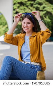 A cute smiling young woman in casual wear sitting at the park. Positive emotions concept, a fashionable girl with long dark hair touching her blue headband.
