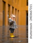 A cute stylish two-year-old toddler kid girl in a gray coat and yellow boots walks outdoors near buildings with glass walls