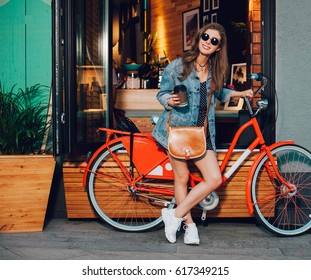 Cute surfer girl in a summer dress, denim jacket, sunglasses and bag stands with red vintage bicycle in LA city, California. Sunny summer. On background surf cafe. Outdoor.