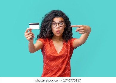 Cute surprised dark skinned girl in trendy spactacles pointing at credit card in her hand with index finger with wide-open eyes face expression.